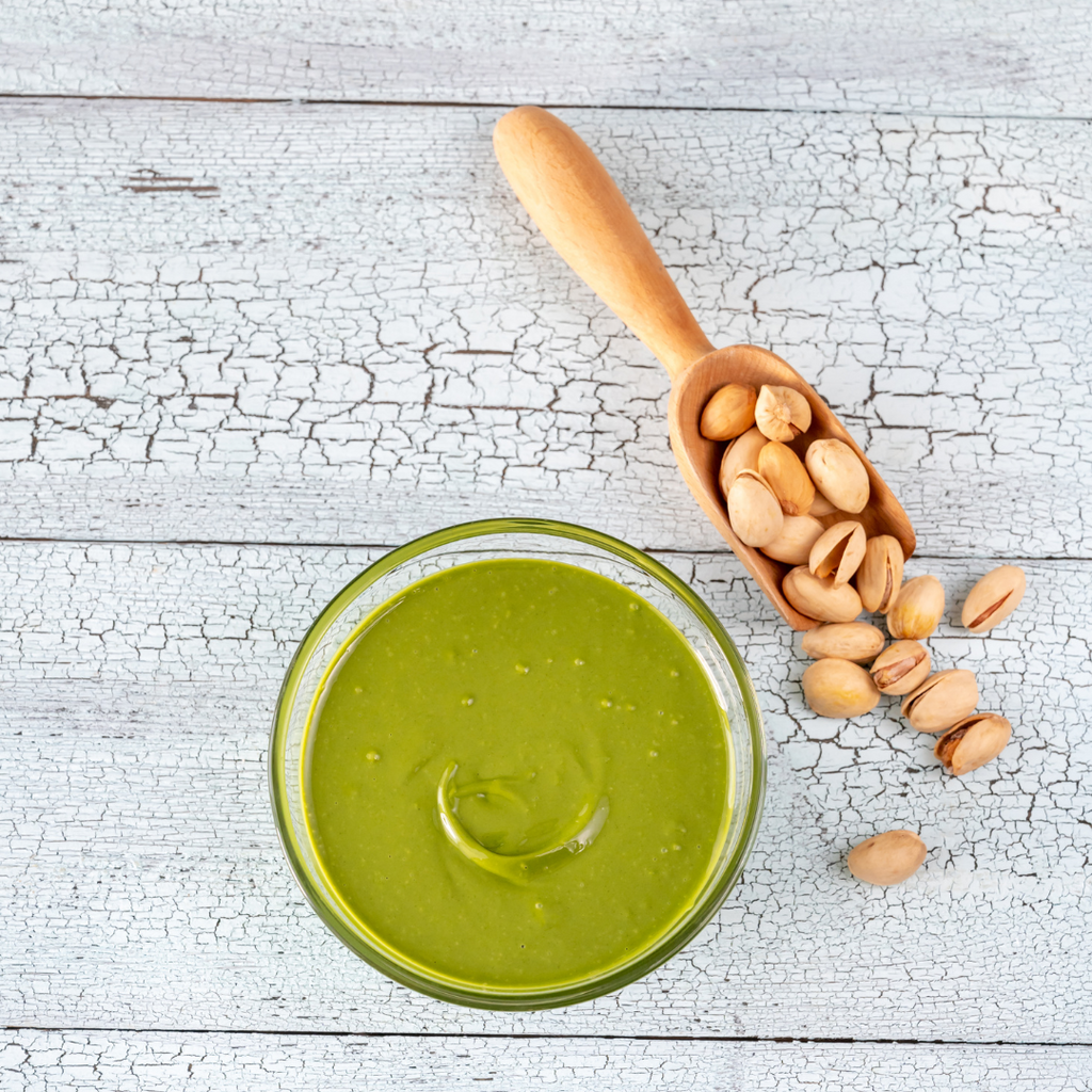 A bowl of pistachio nut butter, complemented by some loose pistachio nuts, presented on a white wooden table.