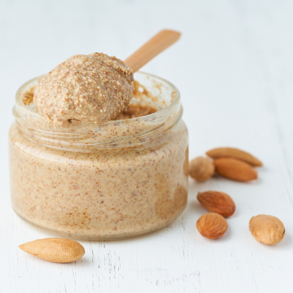 A jar of natural almond butter accompanied by a wooden spoon, placed on a brown wooden table.