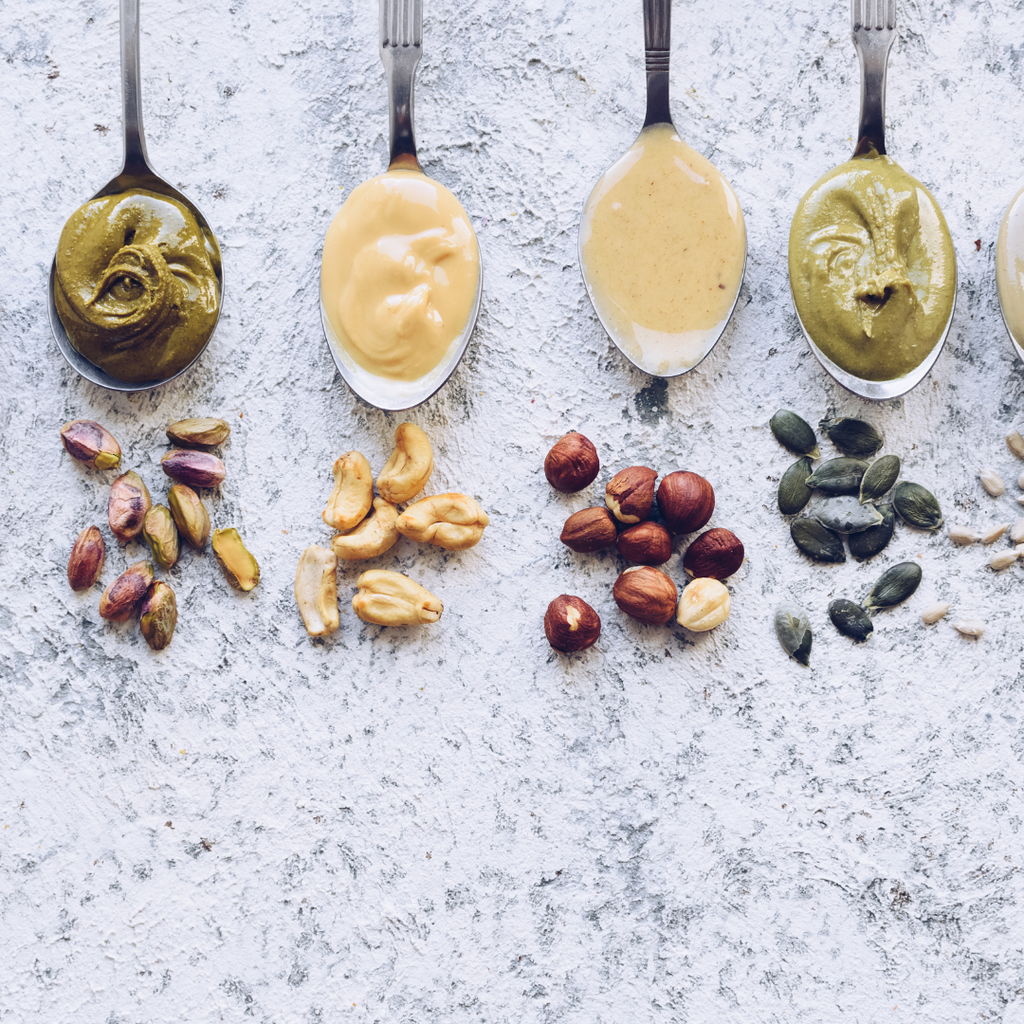 Assorted nut butters on spoons including peanut, hazelnut, pistachio and pumpkin seed, neatly arranged.