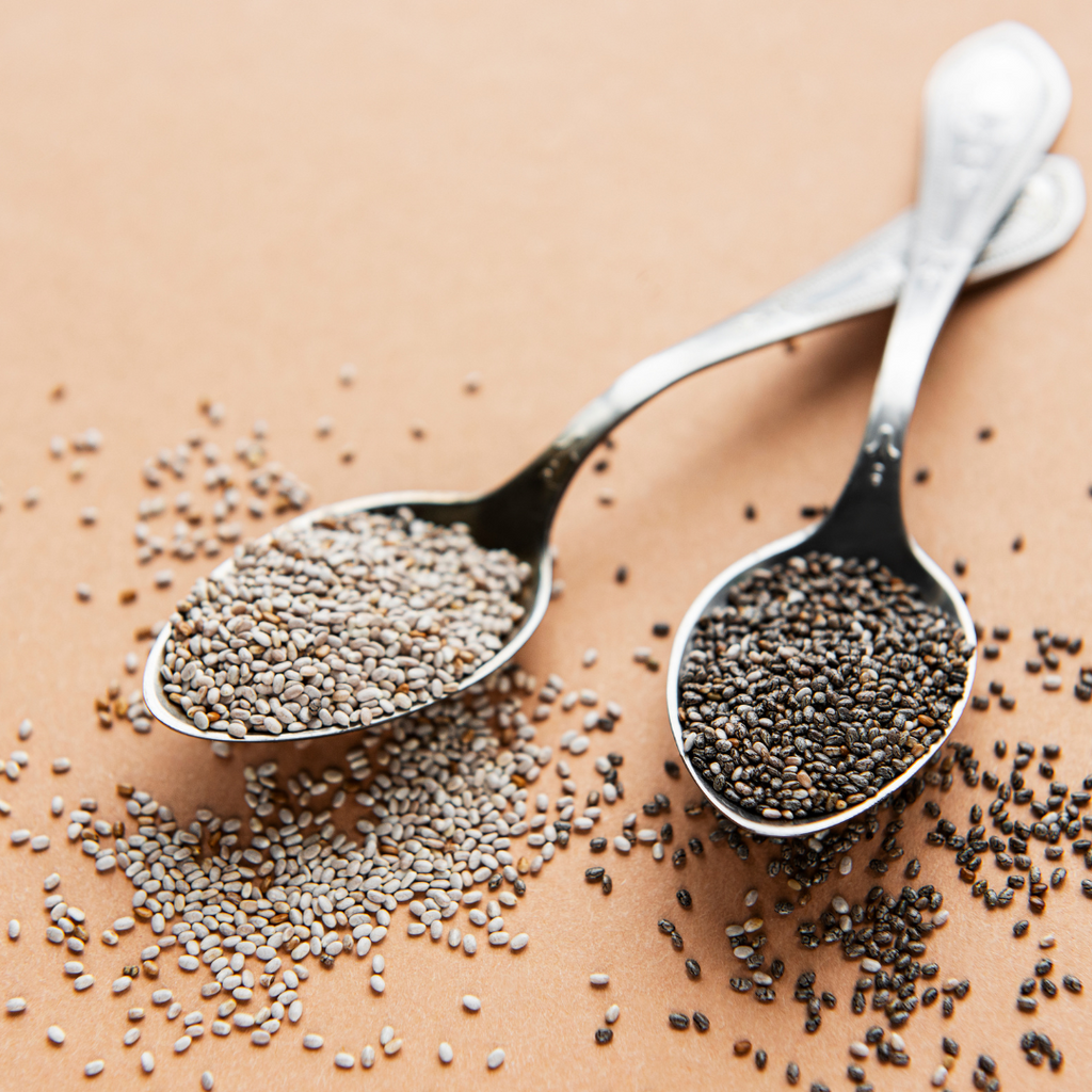 2 silver spoons rested on a table, one containing white chia seeds and one with black chia seeds.