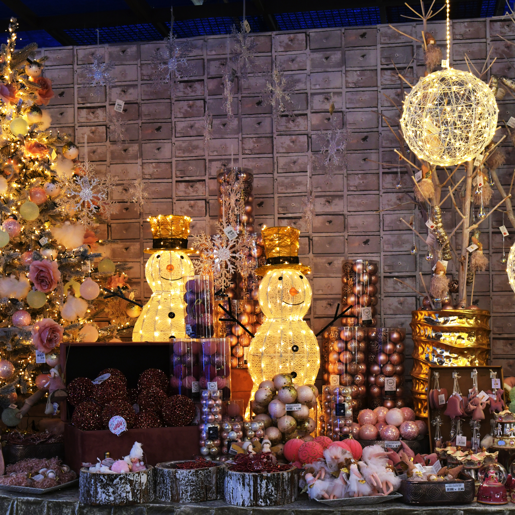 A display of festive decorations featuring two glowing snowmen with golden top hats, surrounded by Christmas ornaments and decorative lights. A decorated tree and other holiday items complete the cozy and colorful scene.