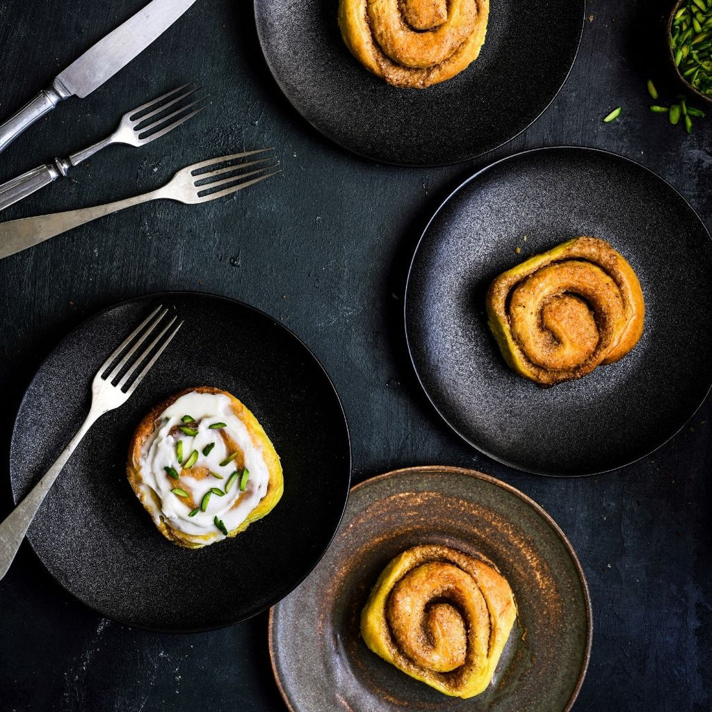 Four black plates, each with a saffron pistachio roll, one with cream cheese frosting and chopped pistachios, set on a dark surface with cutlery.