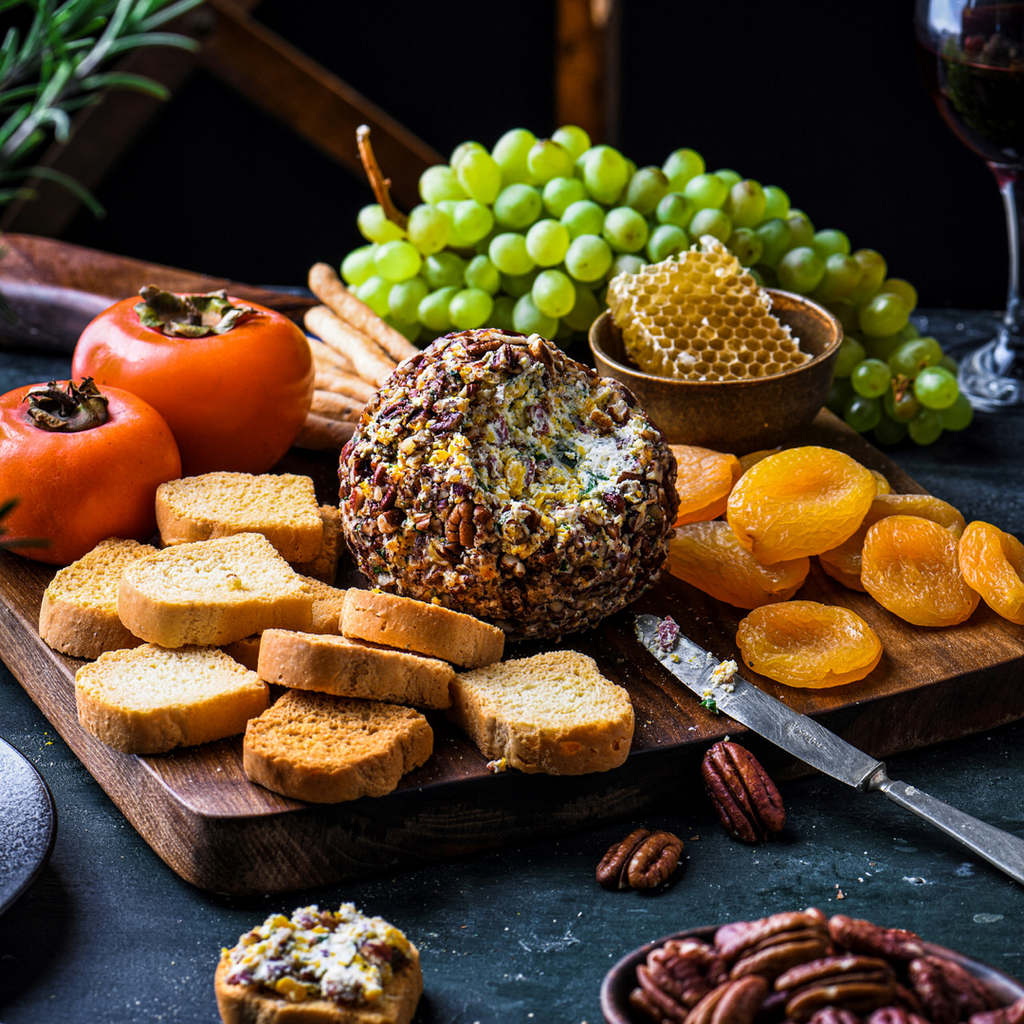 Knife slicing into a bacon pecan cheeseball in the middle of a wooden board surrounded by crostini, grapes, tomato, elegant apricots and a piece of honeycomb in a bowl