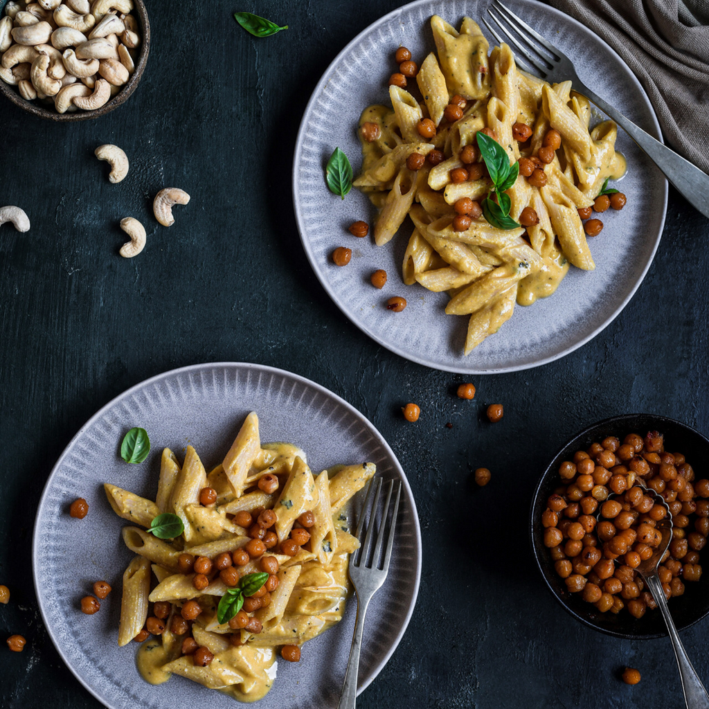 Two plates of creamy cashew pasta topped with crispy roasted chickpeas and garnished with fresh basil leaves, with scattered cashews and chickpeas on a dark background.