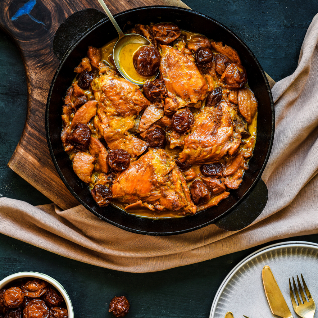 A skillet with braised chicken thighs and prunes, served in a rich golden-brown sauce, set on a wooden cutting board with a beige napkin.