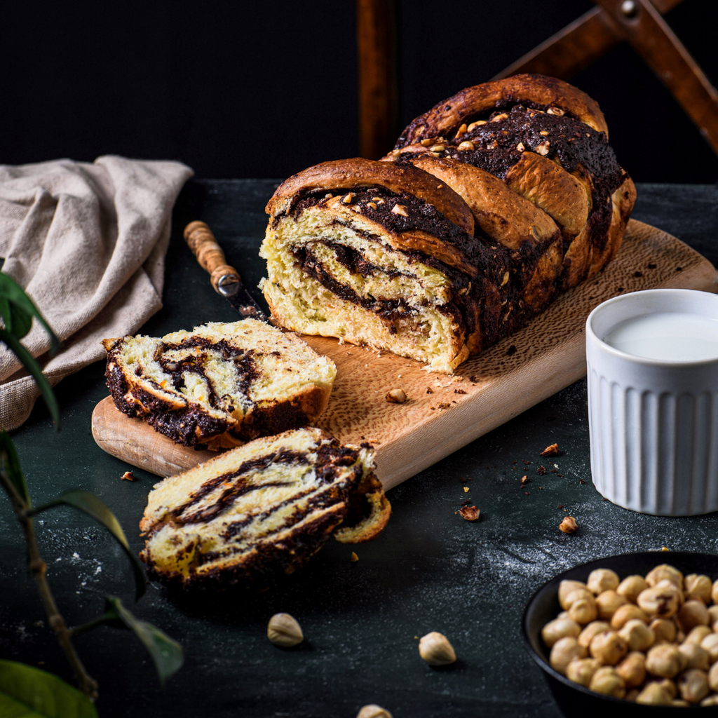 Sliced chocolate hazelnut babka on a wooden bread board on a black table next to ceramic white mug filled with milk.