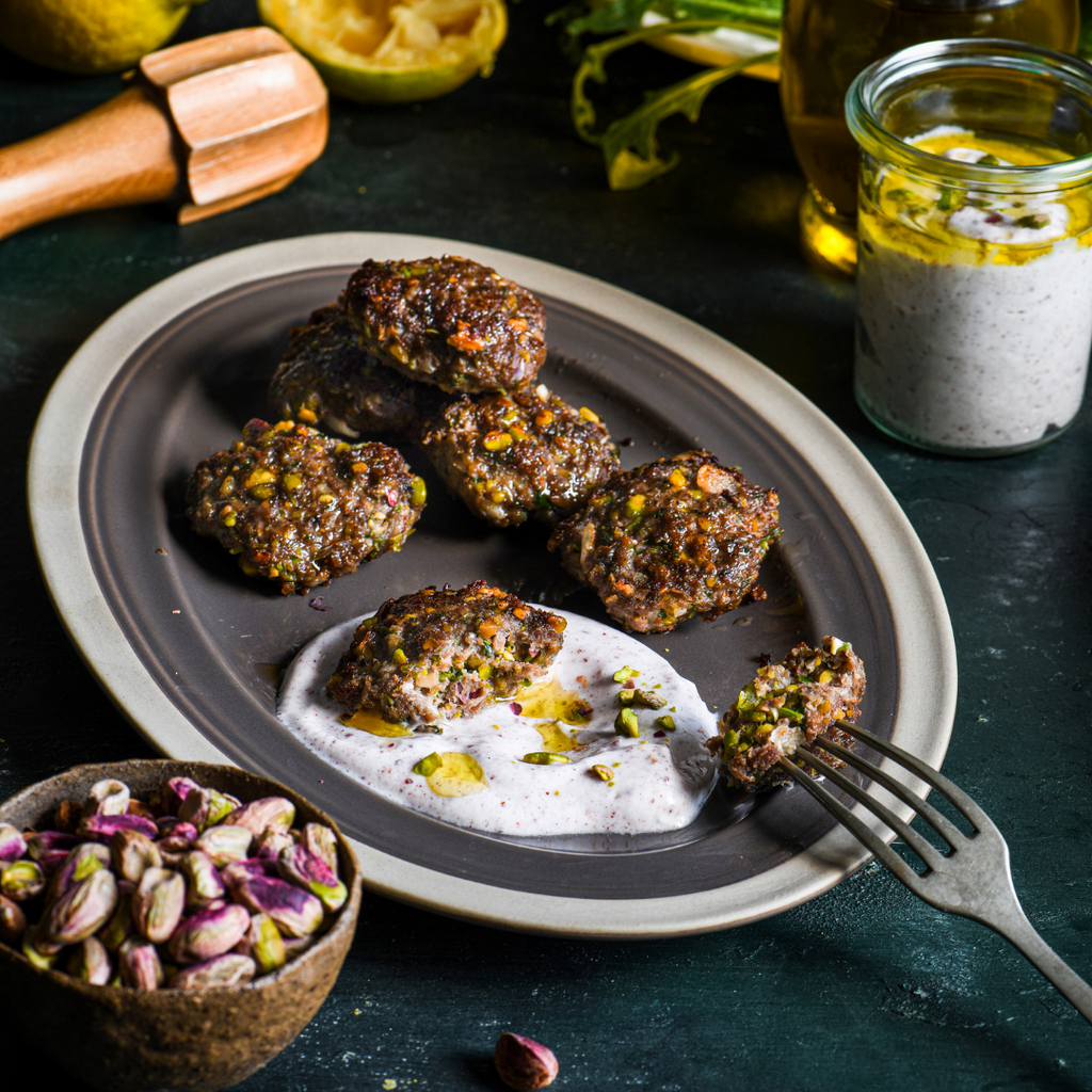 lamb pistachio patties with a side of greek yogurt sauce on a plate next to bowl of shelled pistachios 
