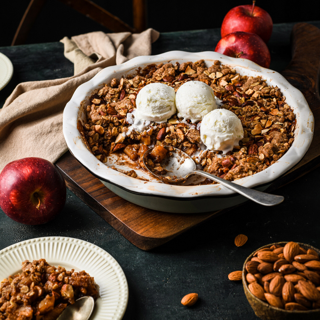 Pie dish containing almond apple crisp dessert and three scoops of ice cream on top surrounded by red apples and a bowl of whole almonds