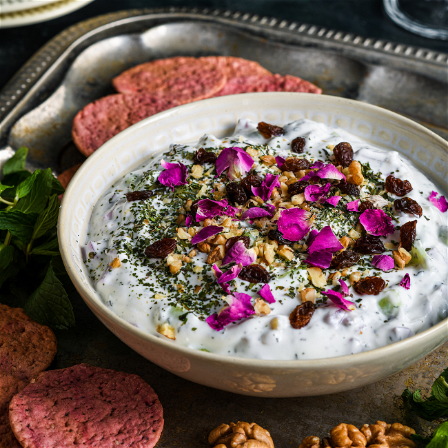 A close-up of a Persian-style yogurt and cucumber dip in a white bowl, garnished with dried rose petals, raisins, chopped walnuts, and dried herbs. 