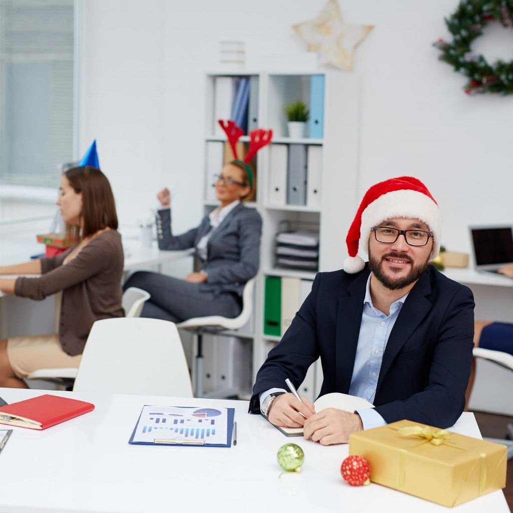 Smiling man in a suit and Santa hat sits at a decorated office desk.