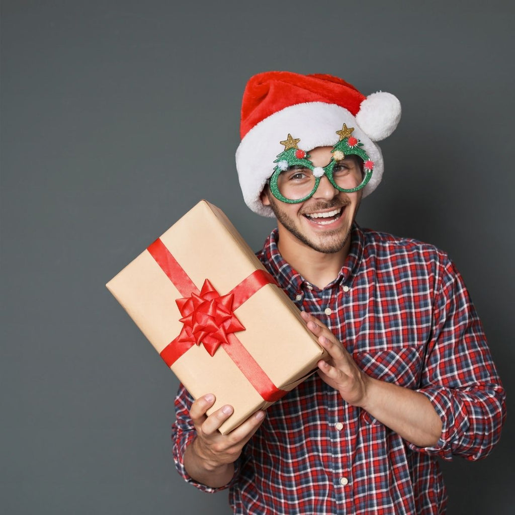 Smiling man in a Santa hat and festive glasses holding a wrapped gift with a red bow.