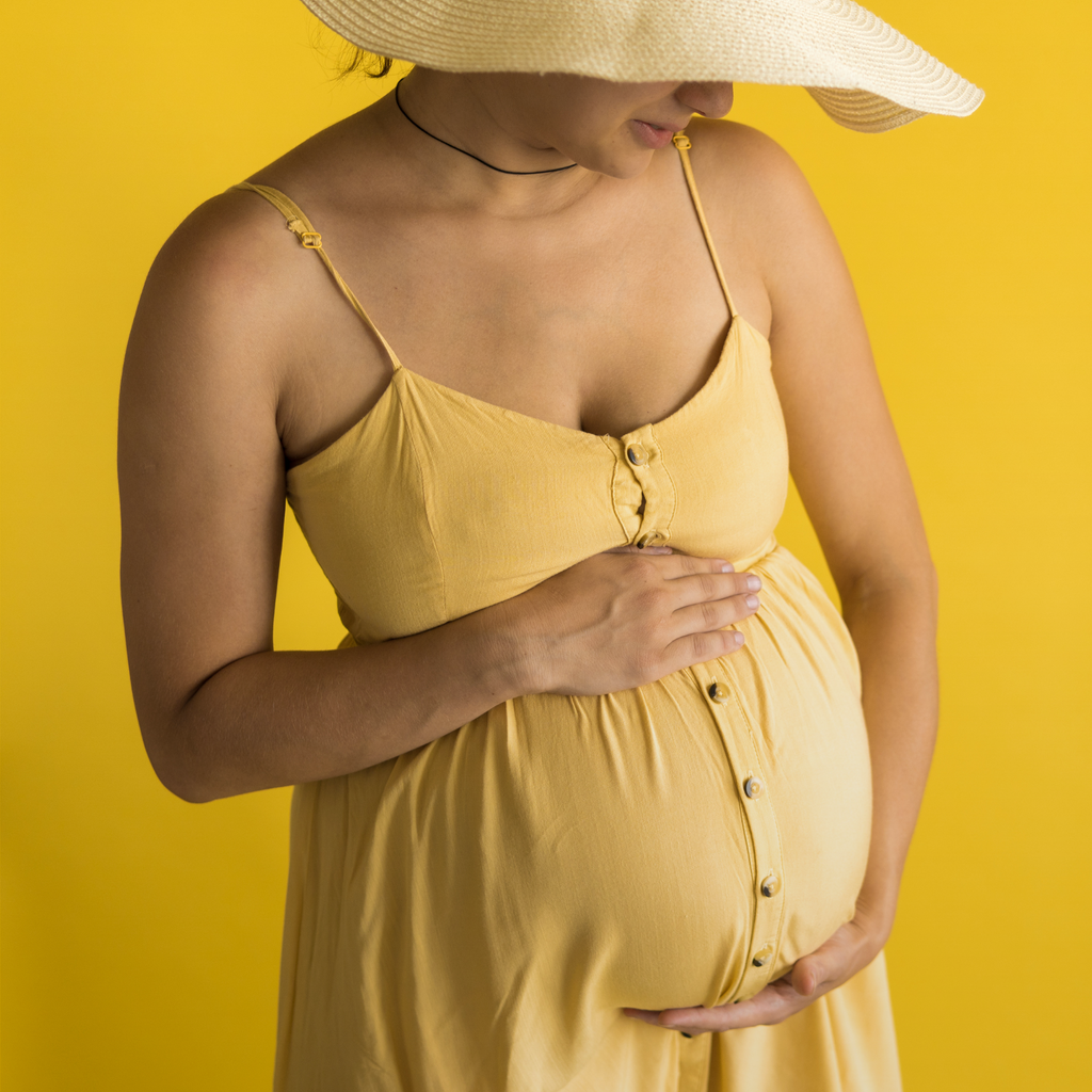 A pregnant woman in a yellow dress cradling her belly with both hands, standing against a yellow background.
