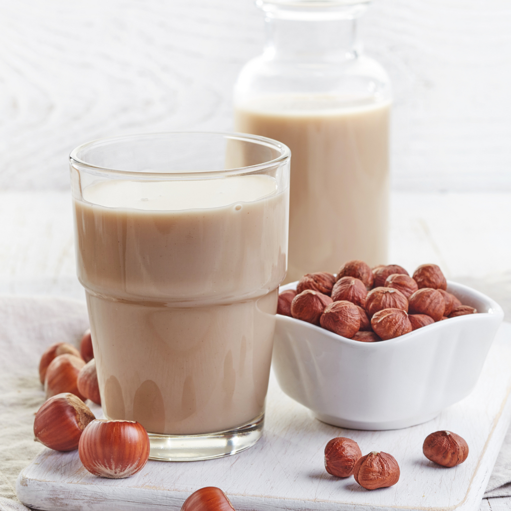 A glass of homemade hazelnut coffee on a white table, next to a bowl of whole, unshelled raw hazelnuts.