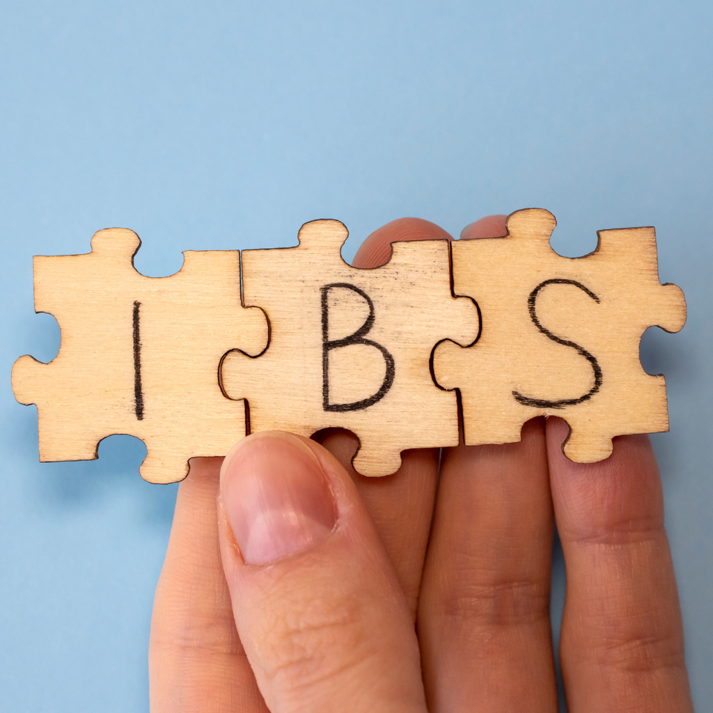 Wooden puzzle pieces spelling "IBS" held by fingers against a blue background.