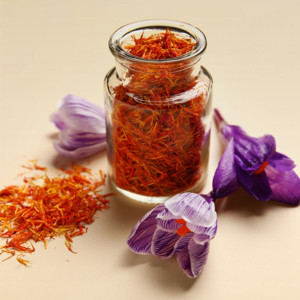 A glass jar filled with red saffron threads sits beside two purple crocus flowers and a small pile of loose saffron on a beige surface.