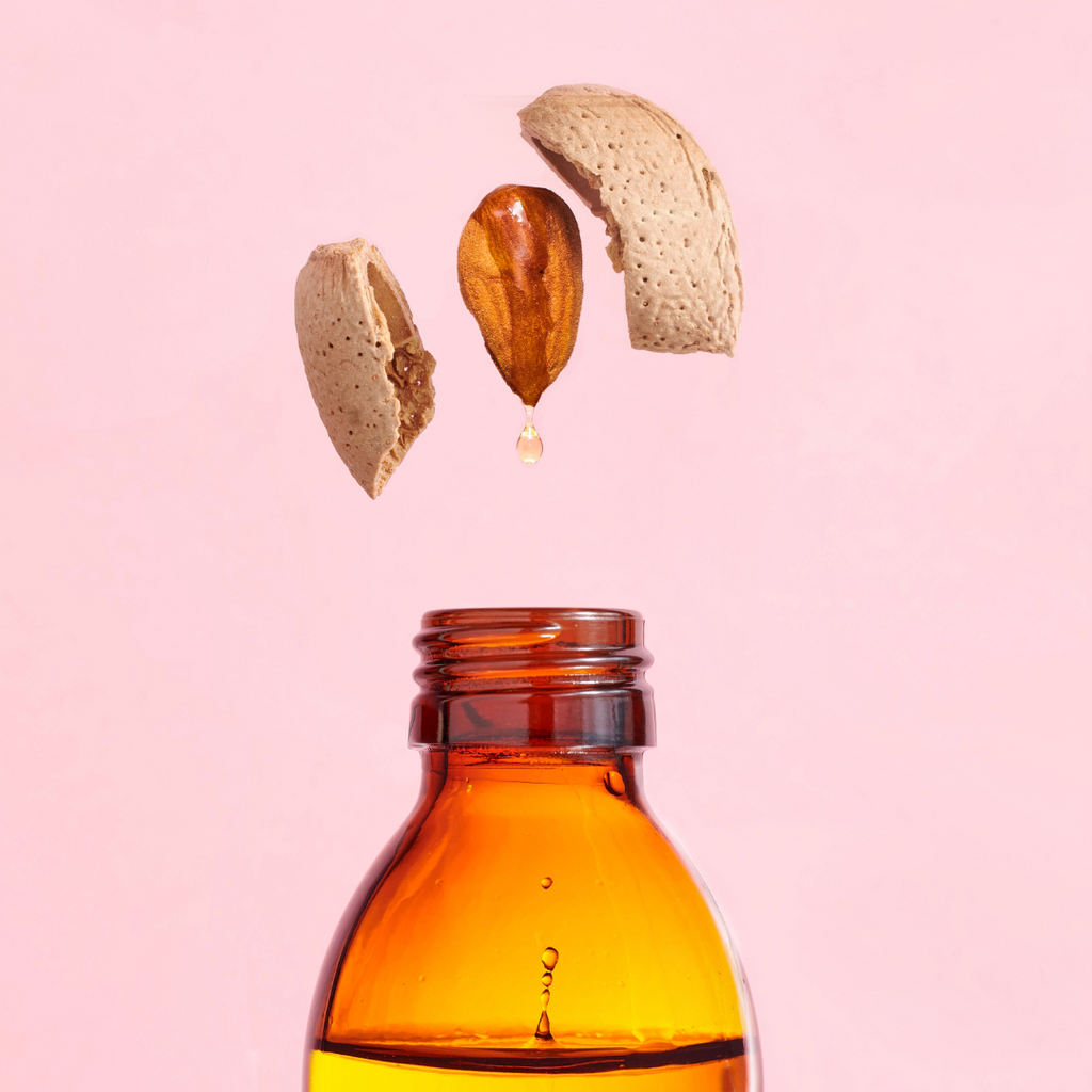 An almond shell breaking open over a bottle of almond oil: a clear, light-textured oil derived from almonds. Used in skincare, haircare, and cooking.