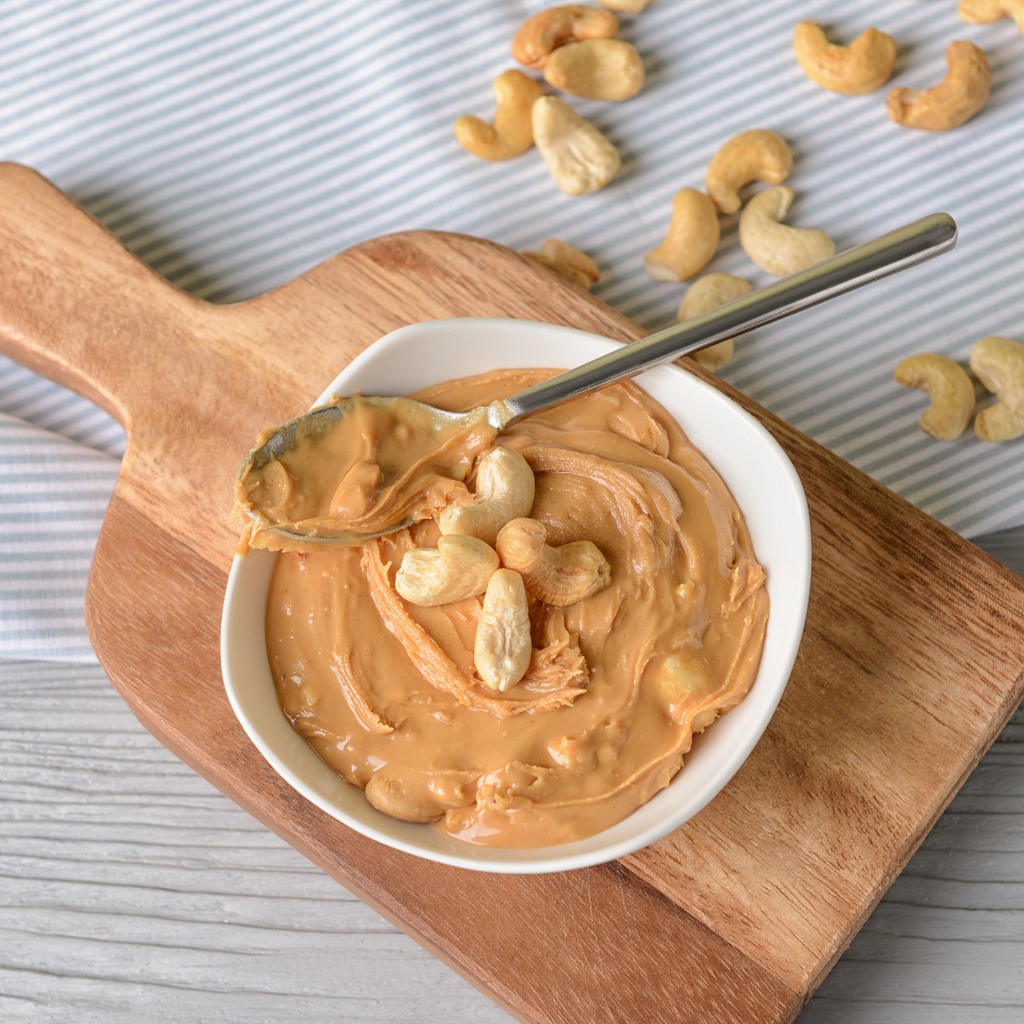 A bowl of cashew nut butter placed on a wooden serving board next to some loose scattered cashews.