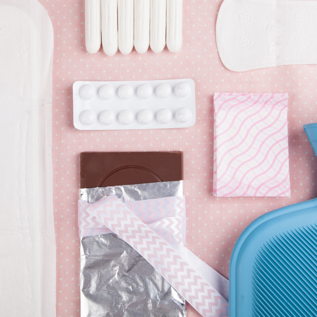 Flat lay of period care essentials, including a chocolate bar, a hot water bottle, pain relief tablets, and sanitary products, arranged on a soft pink background to highlight comfort and nourishment during menstrual cycles.